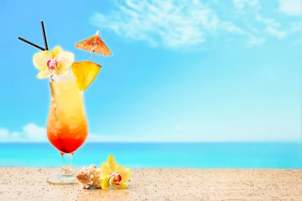 Summer cocktail Tequila Sunrise with tropical flowers,  blured sea and sky on background. Summer holidays and relax concept.