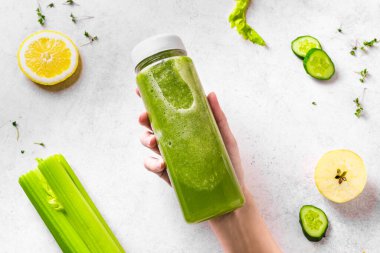 Kadın elinde yeşil meyve püresi şişesi ve beyazın üzerindeki malzemeler, üst manzara. Taze çiğ detoks smoothie veya sağlıklı yemek için yeşil meyve suyu..