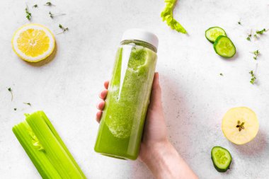 Kadın elinde yeşil meyve püresi şişesi ve beyazın üzerindeki malzemeler, üst manzara. Taze çiğ detoks vejetaryen smoothie veya sağlıklı beslenmek için yeşil meyve suyu..