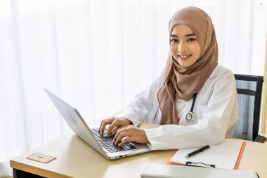 Kendine güvenen Müslüman kadın doktor masasında oturuyor, klinik ofisinde dizüstü bilgisayarla yazıyor, kameraya bakıyor.