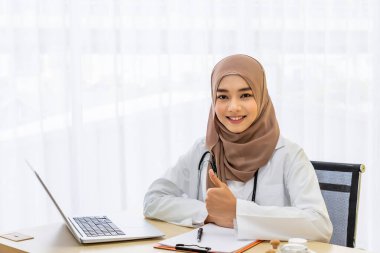 Kendine güvenen Müslüman kadın doktor masasında oturuyor, klinik hastane ofisinde dizüstü bilgisayarla başparmağıyla yazı yazıyor, kameraya bakıyor.