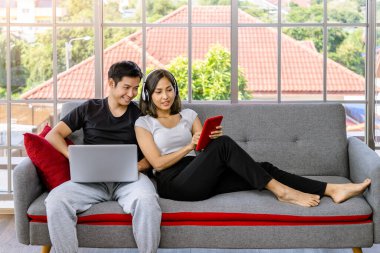 Genç Asyalı çift evde çalışan dizüstü bilgisayar ve dijital tablet kullanarak koltukta rahatlıyor.