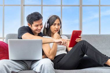 Genç Asyalı çift evde çalışan dizüstü bilgisayar ve dijital tablet kullanarak koltukta rahatlıyor.