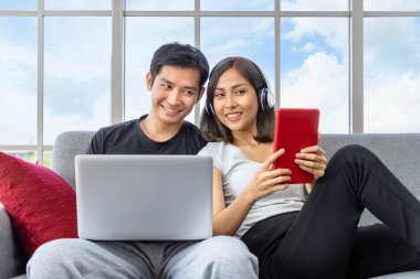 Genç Asyalı çift evde çalışan dizüstü bilgisayar ve dijital tablet kullanarak koltukta rahatlıyor.