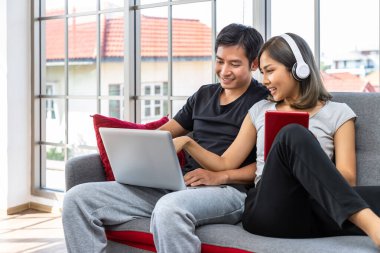 Genç Asyalı çift evde çalışan dizüstü bilgisayar ve dijital tablet kullanarak koltukta rahatlıyor.