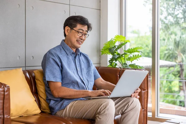 Happy Asian senior retired man sitting on couch sofa, using laptop computer
