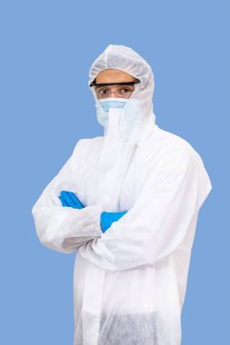 Kişisel Koruyucu Ekipman (PPE), cerrahi maske ve koruma gözlüğü takan kendinden emin Orta Doğulu erkek Arap araştırmacı tıp doktoru portresi açık mavi arka planda izole edilmiş çapraz kollar