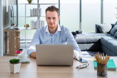 Evde çalışan, oturma odasında dizüstü bilgisayarla oturan ve kameraya bakan gülümseyen beyaz bir adamın portresi.