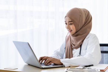 Kendine güvenen Müslüman kadın doktor masasında oturmuş klinik ofisinde dizüstü bilgisayarla yazı yazıyor.