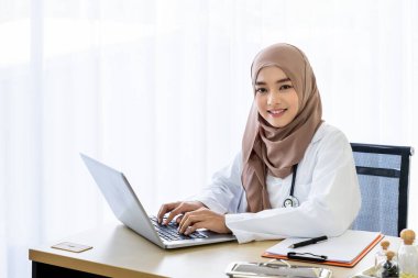 Kendine güvenen Müslüman kadın doktor masasında oturuyor, klinik ofisinde dizüstü bilgisayarla yazıyor, kameraya bakıyor.