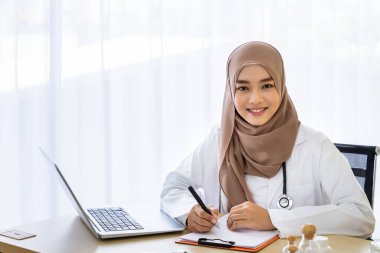 Kendine güvenen Müslüman kadın doktor masasında oturuyor, hastane ofisinde dizüstü bilgisayarla bir şeyler yazıyor, kameraya bakıyor.
