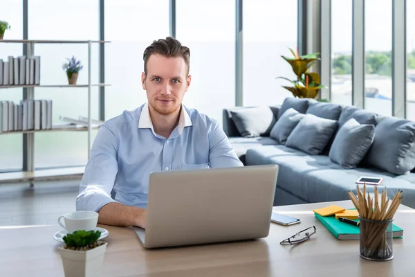 照片中年轻的白人自由职业者在家里工作 坐在客厅的桌子旁 用笔记本电脑打字 看着相机 — 图库照片