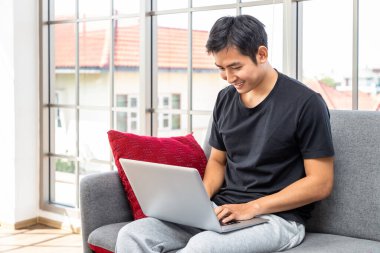 Oturma odasında dizüstü bilgisayar kullanan mutlu Asyalı genç adam ev konseptiyle çalışıyor..