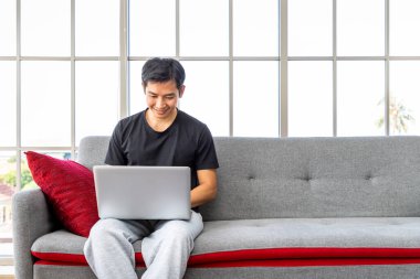 Oturma odasında dizüstü bilgisayar kullanan mutlu Asyalı genç adam ev konseptiyle çalışıyor..
