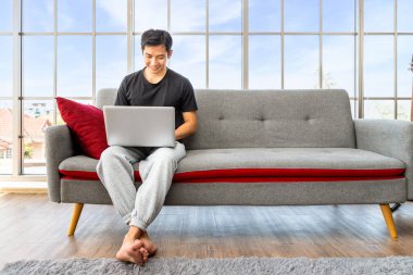 Oturma odasında dizüstü bilgisayar kullanan mutlu Asyalı genç adam ev konseptiyle çalışıyor..