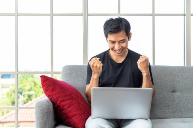 Oturma odasında dizüstü bilgisayar kullanan mutlu bir Asyalı. Başarı için yumruğunu kaldır. Ev kavramından çalış..
