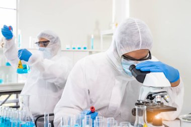 Laboratuvar kişisel koruyucu ekipman giysisi araştırmacısı veya PPE aşı, bilim ve tıbbi konsepti keşfetmek için mikroskop kullanarak biyolojik tehlike virüsü üzerinde deney yapıyor..