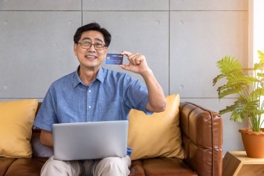 Kıdemli Asyalı adam kanepeye oturmuş bilgisayarını kullanıyor. İnternetten alışveriş ve ödeme için kredi kartı tutuyor. Kameraya bakıyor.