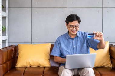 Kıdemli Asyalı adam kanepeye oturmuş bilgisayarını kullanarak online alışveriş ve ödeme için kredi kartı tutuyor.