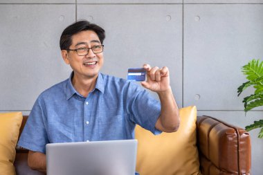 Kıdemli Asyalı adam kanepeye oturmuş bilgisayarını kullanarak online alışveriş ve ödeme için kredi kartı tutuyor.