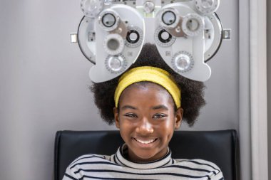 Afrikalı genç bir kız klinikte ya da optik dükkanda göz testi yapıyor. Foropter ekipmanında oturuyor. Göz konsepti.