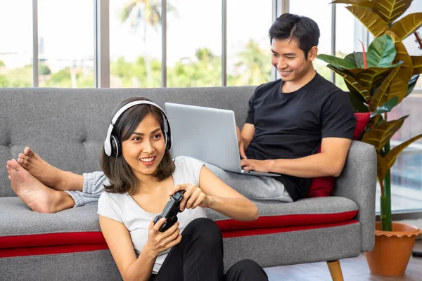 Young Asian couple in living room, man sitting on sofa using laptop computer working from home, and woman playing game with gamepad and headphone