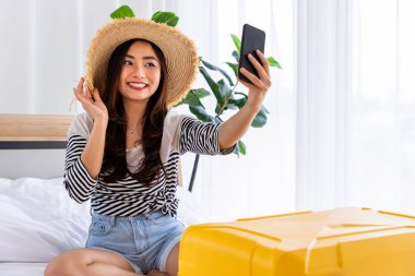 Mutlu Asyalı kadın seyahat için bavul hazırlıyor, yatak odasında oturuyor ve selfie çekiyor.