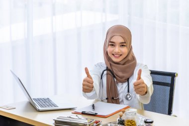 Kendine güvenen Müslüman kadın doktor masasında oturuyor, klinik hastane ofisinde dizüstü bilgisayarla başparmağıyla yazı yazıyor, kameraya bakıyor.