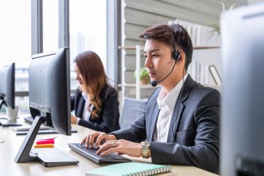 Genç Asyalı erkek müşteri hizmetleri destek operatörü ekip çağrı merkezinde kulaklıklar ve bilgisayarla çalışıyor..