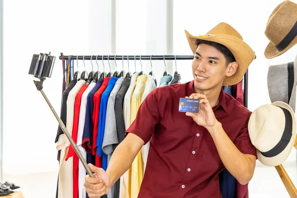 Young Asian male online merchant blogger using mobile phone camera with selfie stick to live video to review and sell product, showing credit card. Social media, Influencer, online shopping, and payment concept.