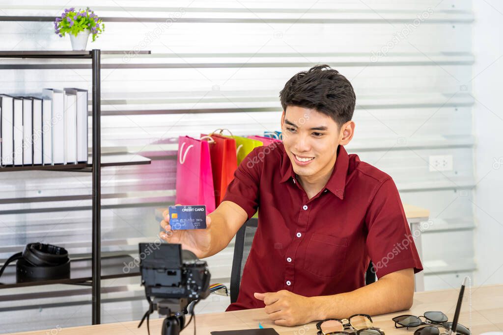 Young Asian male online merchant blogger using camera to live video to review and sell product, showing credit card. Social media, Influencer, online shopping, and payment concept.