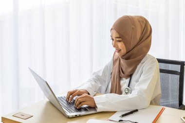 Kendine güvenen Müslüman kadın doktor masasında oturmuş klinik ofisinde dizüstü bilgisayarla yazı yazıyor.