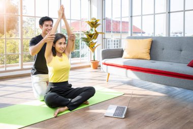 Çekici genç Asyalı çift evdeki oturma odasında yoga yapıyor, online ders izlemek için dizüstü bilgisayar kullanıyorlar.