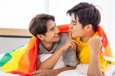 Mutlu Asyalı eşcinsel çift yatakta uzanıyor ve parmaklarıyla dokunuyor gökkuşağı gurur bayrağı, LGBT konsepti.