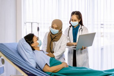 Yataktaki yaşlı kadın hastayı ziyaret eden ve onunla ilgilenen bir grup doktor kalp atışlarını steteskopla kontrol ediyor ve dizüstü bilgisayar kullanıyor..