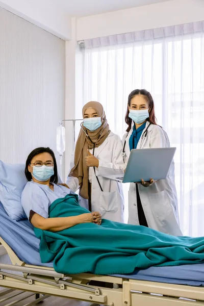 Team of doctors visiting and taking care of senior female patient in bed, with stethoscope and laptop computer, looking at camera with thumb-up.