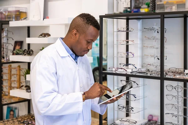 Portrait of African optician in optical shop store, using digital tablet to count and check eyeglasses stock. Eyecare and shopkeeper concept.