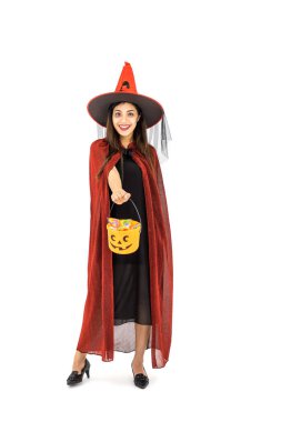 Cadılar Bayramı kostümü giymiş Asyalı bir kadın kırmızı pelerinli, beyaz arka planda, şeker ya da şaka için turuncu şeker kovası tutuyor, kameraya bakıyor.