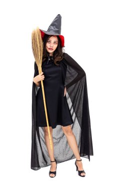 Siyah pelerinli cadı kostümü giymiş Asyalı bir kadın beyaz arka planda elinde süpürge sopasıyla kameraya bakıyor.