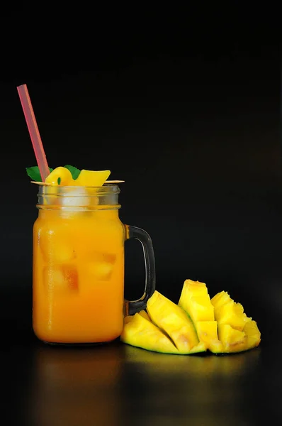 A glass mug of yellow fruit nectar with ice and a straw stands next to a cut mango on a black background. Close-up.