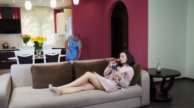 Bir adam ev işi yapıyor, genç bir kız kanepede dinleniyor, akıllı telefona bakıyor ve kırmızı şarap içiyor..