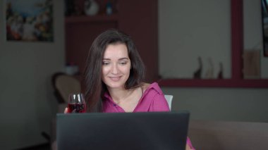 İzole edilmiş bir kız dizüstü bilgisayarında oturup bardaktan kırmızı şarap içiyor, sohbet ediyor, gülümsüyor ve gülüyor..