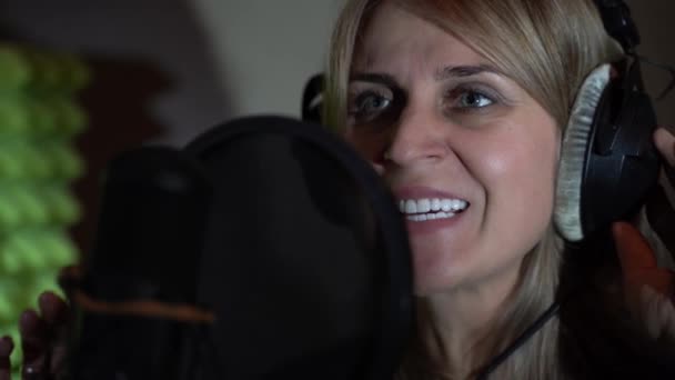 Una donna in un piccolo studio di registrazione canta in un microfono indossando cuffie nere davanti a un microfono da studio. Una donna canta una canzone in uno studio di registrazione. — Video Stock