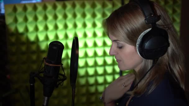 Una donna in un piccolo studio di registrazione canta in un microfono indossando cuffie nere davanti a un microfono da studio. Una donna canta una canzone in uno studio di registrazione. — Video Stock