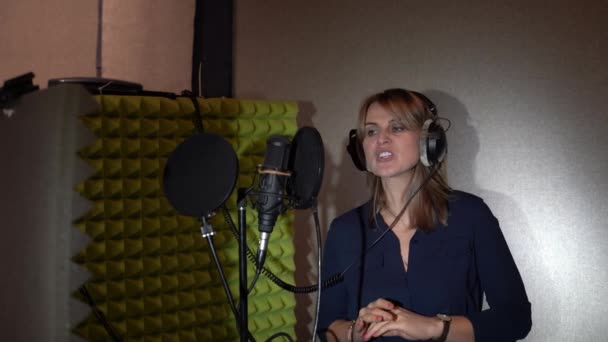 Женщина в маленькой студии звукозаписи поет в микрофон в черных наушниках перед студийным микрофоном. Женщина поет песню в студии звукозаписи. — стоковое видео