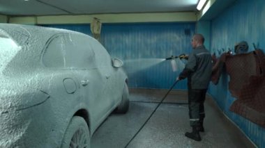 Beyaz bir adam araba yıkamada çalışır. Bir araba yıkama işçisi arabayı yıkar. Bir servis çalışanı yıkama için arabaya beyaz köpük döker.