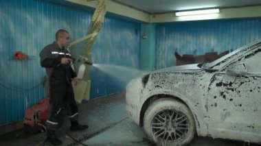 Beyaz bir adam araba yıkamada çalışır. Bir araba yıkama işçisi arabayı yıkar. Bir servis çalışanı yıkama için arabaya beyaz köpük döker.