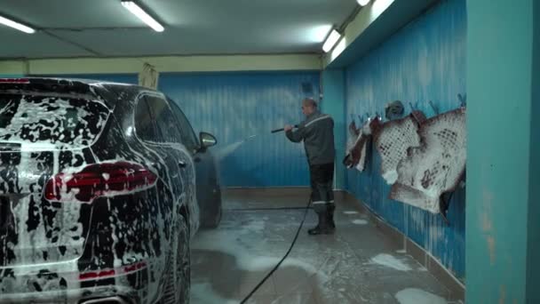 Un hombre caucásico trabaja en un lavado de autos, un trabajador de lavado de autos lava el auto. El empleado de servicio lava la espuma de la máquina con una fuerte presión de agua — Vídeos de Stock