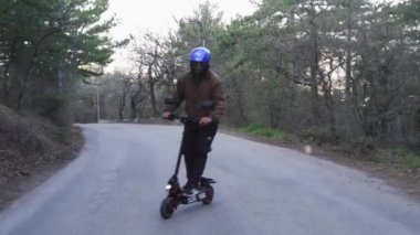 Koruyucu kask takan ve güçlü bir elektrikli scooter kullanan bir sürücü orman yolunda araba sürer. Adamın biri ormanda beton bir yolda elektrikli scooter sürmeye başlıyor..