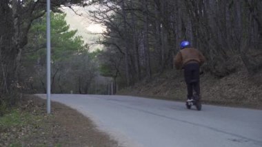 Koruyucu kask takan ve güçlü bir elektrikli scooter kullanan bir sürücü orman yolunda araba sürer. Ormanda elektrikli scooterlı bir adam beton bir yolda hızla kameradan uzaklaşıyor.